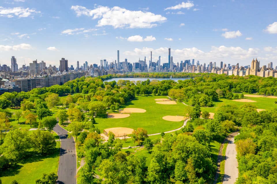 Aerial view of Central Park in New York City, surrounded by green trees and with the Manhattan skyline in the background under a bright blue sky.